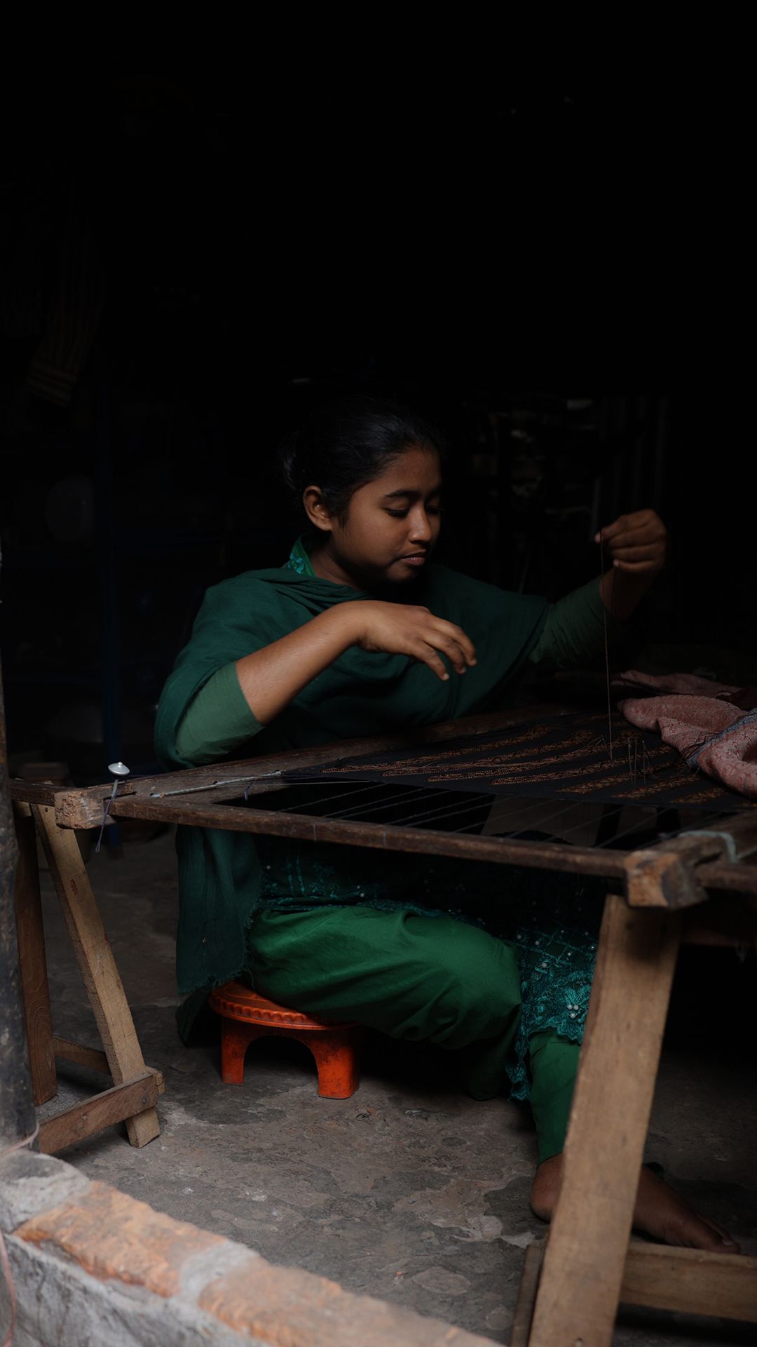 A woman sits in a dark room weaving cloth on a large loom.